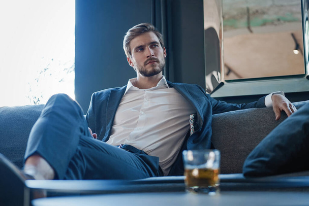 Young and charming businessman is thinking about business while sitting on the sofa in his modern office. - 写真・画像