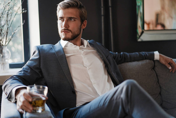 Handsome and successful businessman in stylish suit holding glass whiskey while sitting at office. - 写真・画像