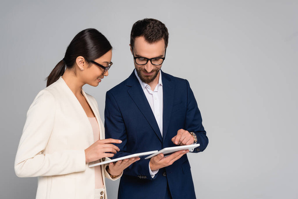 pareja interracial de empresarios en ropa formal y gafas con tabletas digitales aisladas en gris - Foto, Imagen