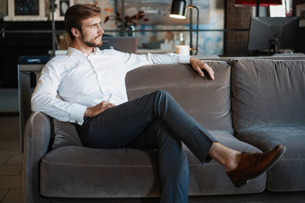 Young and charming businessman is thinking about business while sitting on the sofa in his modern office. - Foto, immagini