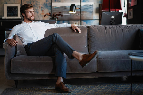 Young and charming businessman is thinking about business while sitting on the sofa in his modern office. - Foto, Bild