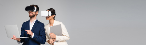 horizontální obrázek podnikatele ukazující prstem na notebook při použití vr sluchátek spolu s kolegou izolovaným na šedé - Fotografie, Obrázek