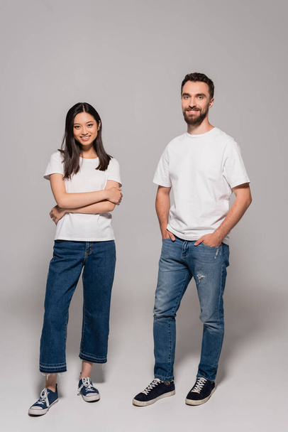 stylish interracial couple in white t-shirts, jeans and gumshoes posing with hands in pockets and crossed arms on grey - Fotografie, Obrázek