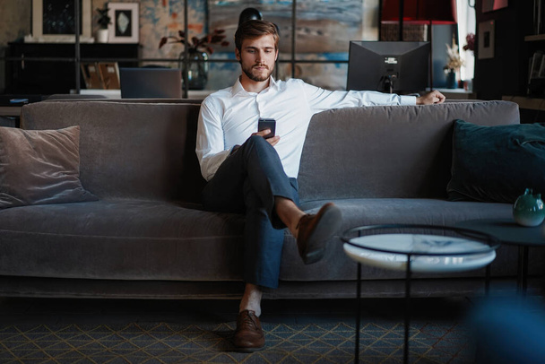 Smiling bearded man making text message on smartphone while sitting sofa at modern office. - Valokuva, kuva