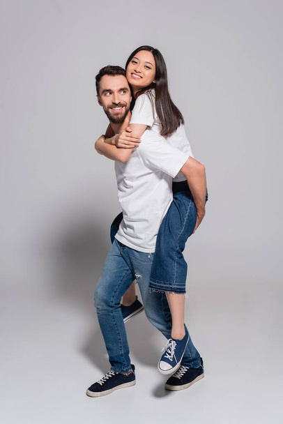 bearded man in white t-shirt and jeans piggybacking asian girlfriend on grey - Photo, Image