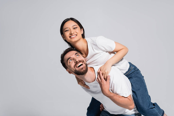 excited man in white t-shirt piggybacking asian girlfriend isolated on grey - Фото, изображение