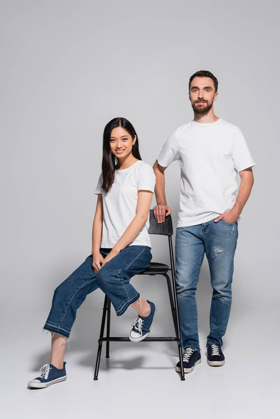 brunette asian woman in white t-shirt and jeans sitting on chair near stylish boyfriend standing with hand in pocket on grey - Fotografie, Obrázek