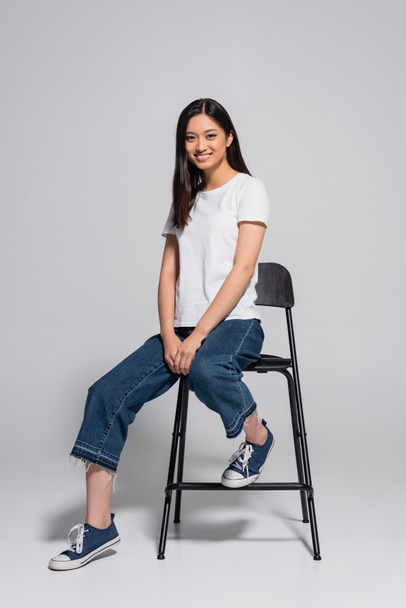 morena mujer asiática en jeans, zapatos de goma y camiseta blanca sentado en la silla y mirando a la cámara en gris - Foto, imagen