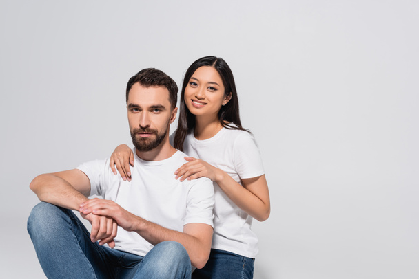 brunette asian woman touching boyfriend and looking at camera while sitting together isolated on white - Foto, imagen