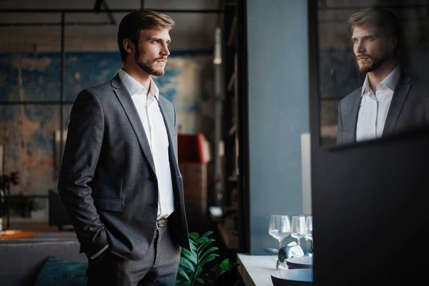 Stylish young bearded man in a suit is standing near the window and looking far - Photo, image