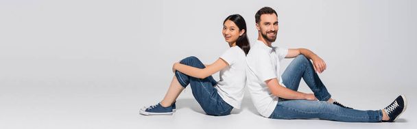 panoramisches Konzept eines stilvollen gemischtrassigen Paares in weißen T-Shirts, Jeans und Gummischuhen, das Rücken an Rücken auf Weiß sitzt  - Foto, Bild