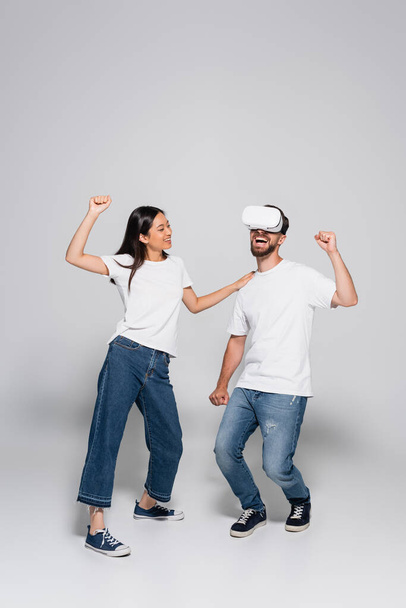 asian woman in jeans and white t-shirt touching shoulder of excited man dancing and singing in vr headset on grey - Foto, imagen