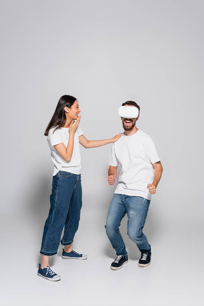 excité homme dans vr casque dansant et chantant près asiatique femme couvrant bouche avec main sur gris - Photo, image