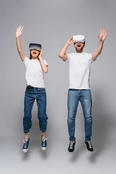 excited couple in vr headsets, jeans and white t-shirts waving hands while levitating in vr headsets on grey - Fotografie, Obrázek