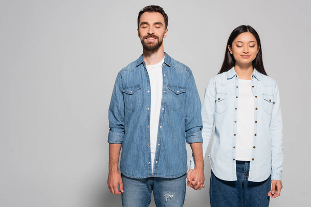 stylish interracial couple in denim clothes and gumshoes holding hands while standing with closed eyes on grey - Fotografie, Obrázek