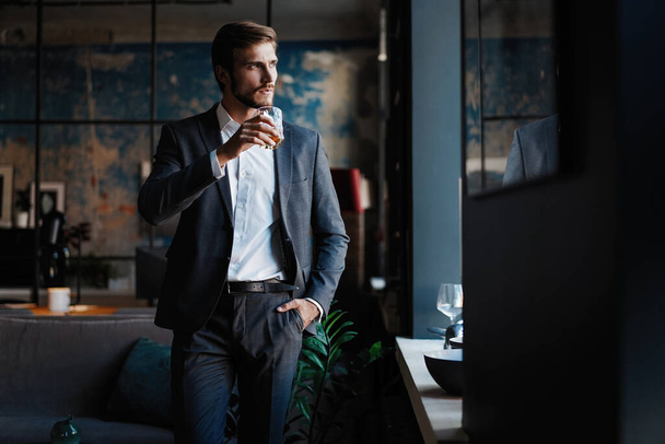 Young handsome businessman stand and hold glass of whiskey in his own office. - Foto, immagini