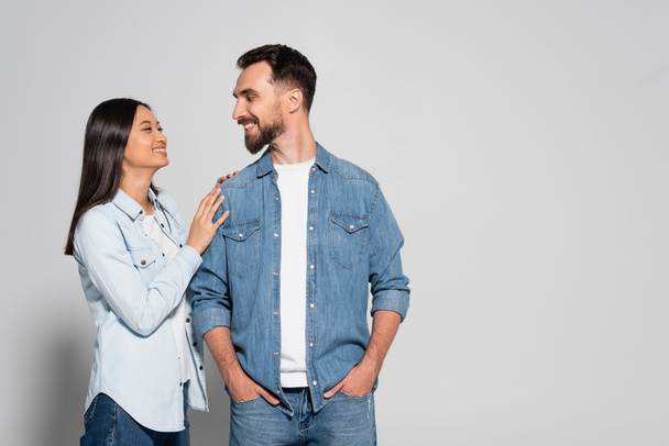 young asian woman touching bearded boyfriend standing with hands in pockets on grey - Fotó, kép