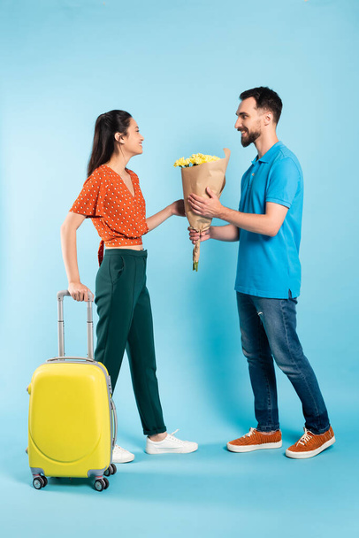 man in polo t-shirt and jeans presenting bouquet to asian woman with yellow suitcase on blue - Foto, Imagem
