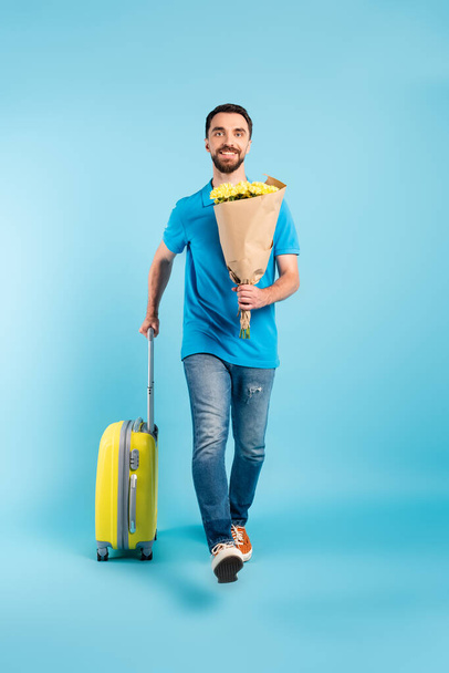 joven turista en camiseta de polo y jeans sosteniendo ramo amarillo mientras camina con la maleta en azul - Foto, Imagen