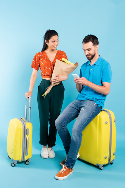 asian woman in red blouse holding bouquet near boyfriend sitting on suitcase with smartphone on blue - Valokuva, kuva