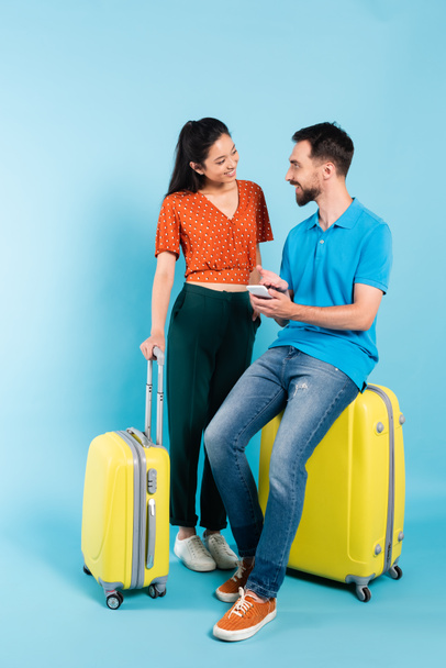 asian woman in red blouse looking at boyfriend sitting on suitcase and pointing with hand at smartphone on blue - Foto, immagini
