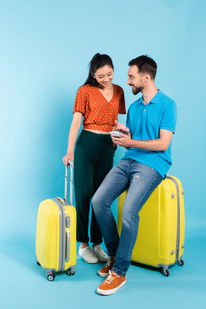 bearded man in polo t-shirt pointing with hand at smartphone while sitting on suitcase near asian girlfriend on blue - Foto, Imagem