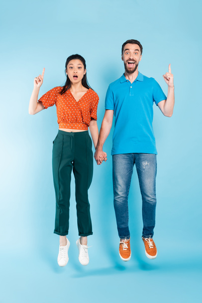 excited asian couple holding hands and showing idea gesture while levitating on blue - Foto, Bild