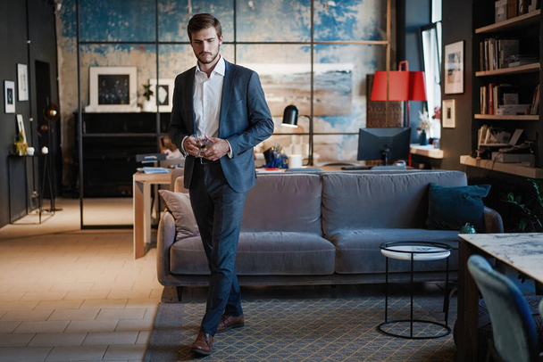Young handsome businessman stand and hold glass of whiskey in his own office. - Foto, Imagem