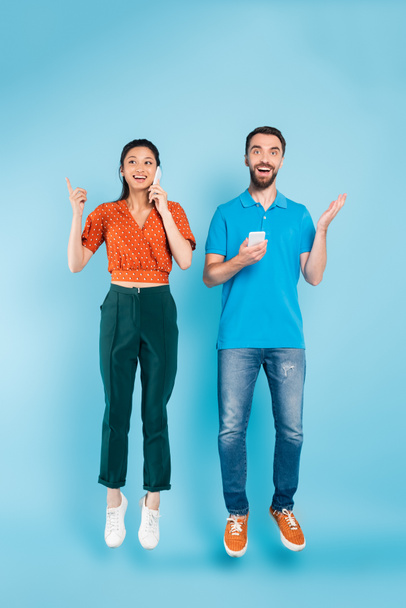 brunette asian woman talking on smartphone and pointing with finger near excited man levitating on blue - Foto, imagen