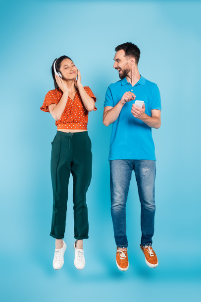 excited man pointing with finger at smartphone near pleased asian woman in headphones levitating on blue - Фото, изображение