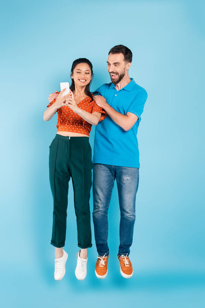 excited man touching shoulders of asian girlfriend using smartphone while levitating on blue - Photo, Image