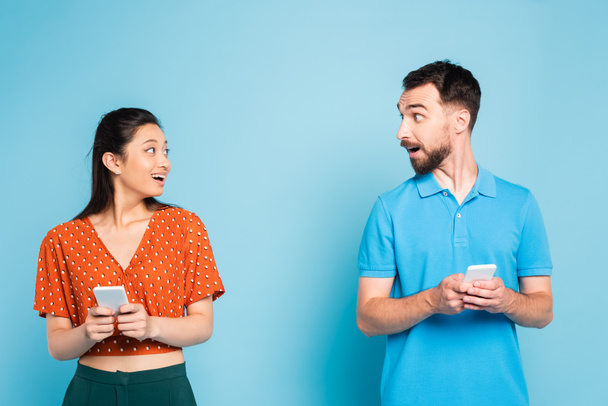 excited interracial couple looking at each other while holding smartphones on blue - Fotó, kép