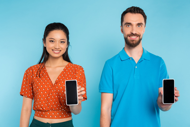 asian woman in red blouse and man in polo t-shirt showing smartphones with blank screen on blue - Foto, afbeelding