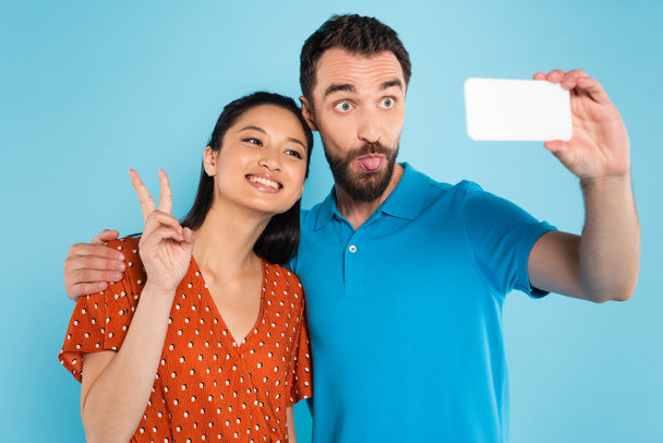 asian woman showing peace sign near boyfriend sticking out tongue while taking selfie isolated on blue - Foto, afbeelding