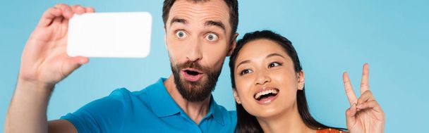 panoramic concept of asian woman showing peace gesture near boyfriend grimacing while taking selfie isolated on blue - Foto, immagini