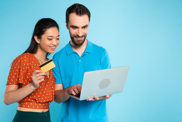 morena asiática mujer en rojo blusa celebración de tarjeta de crédito cerca de hombre en polo camiseta usando portátil en azul - Foto, imagen