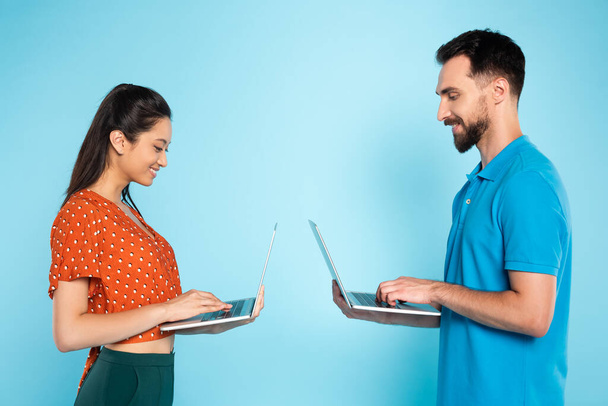 vue latérale de la femme asiatique en chemisier rouge et l'homme en polo t-shirt à l'aide d'ordinateurs portables sur bleu - Photo, image