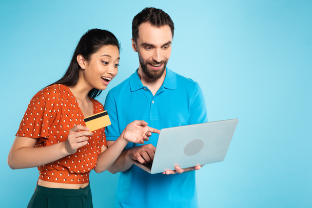 joven hombre utilizando portátil cerca asiático mujer celebración de tarjeta de crédito y señalando con el dedo en azul - Foto, imagen