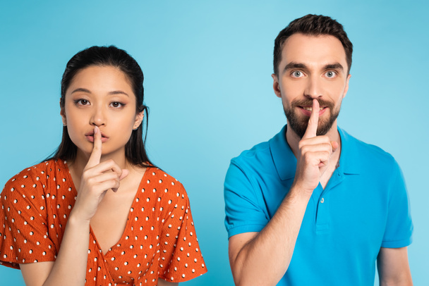 young asian woman in red blouse and man in polo t-shirt showing hush gesture on blue - Foto, Imagem