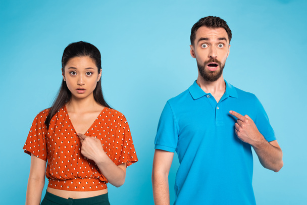 surpris femme asiatique en chemisier rouge et barbu homme en polo t-shirt pointant avec les doigts sur eux-mêmes sur bleu - Photo, image