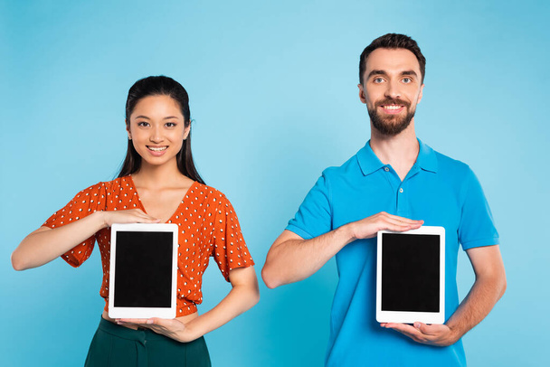 asian woman in red blouse and bearded man in polo t-shirt holding digital tablets with blank screen on blue - Фото, зображення