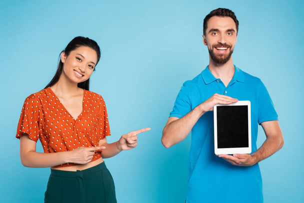 brunette asian woman in red blouse pointing with fingers at digital tablet with blank screen in hands of bearded man on blue - Photo, Image