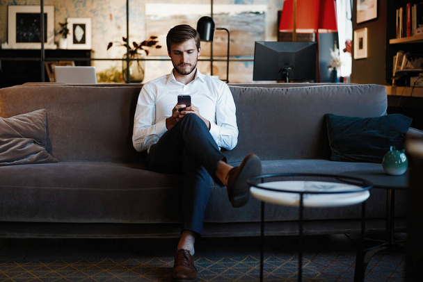 Smiling bearded man making text message on smartphone while sitting sofa at modern office. - Photo, image