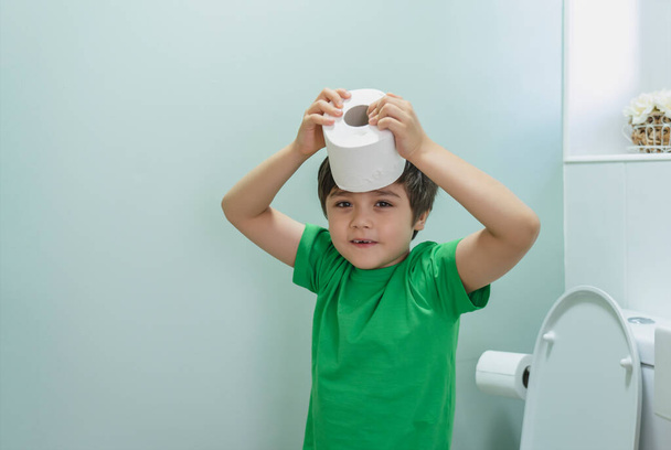 Słodki chłopiec siedzący w toalecie bawiący się papierem toaletowym, chłopiec siedzący w toalecie i zakładający rolkę toalety na głowę, dziecko trzymające białą tkankę, koncepcja opieki zdrowotnej dla dzieci - Zdjęcie, obraz