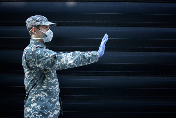 Soldat in Tarnuniform mit Schutzhandschuhen und Maske zeigt Stoppschild mit der Hand vor schwarzem Hintergrund. - Foto, Bild