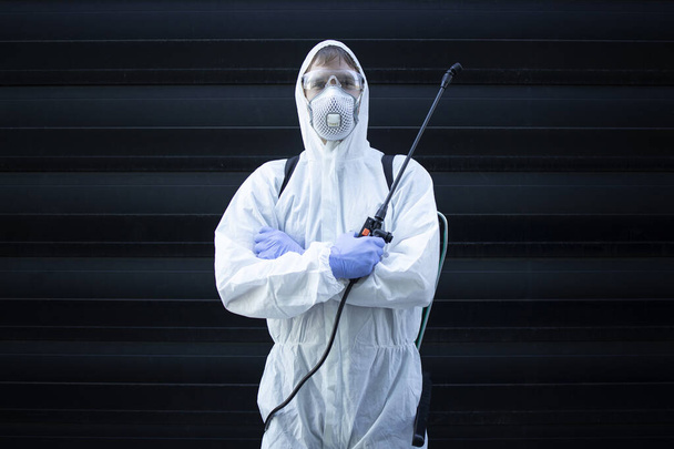 Πορτρέτο του άνδρα σε λευκό κοστούμι χημικής προστασίας κρατώντας ψεκαστήρα με απολυμαντικά χημικά για να σταματήσει την εξάπλωση εξαιρετικά μεταδοτική ιό. Σταματήστε τον κορωνοϊό ή το COVID-19. - Φωτογραφία, εικόνα