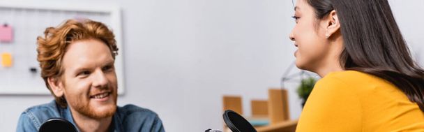 селективное внимание молодого рыжего радиоведущего, интервьюирующего брюнетку азиатку в студии, горизонтальное изображение - Фото, изображение