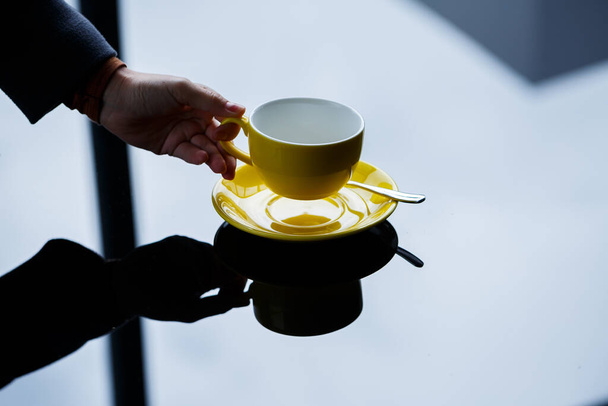Taza amarilla para café o té con un platillo en las manos de una niña en el fondo de una mesa de vidrio. - Foto, imagen