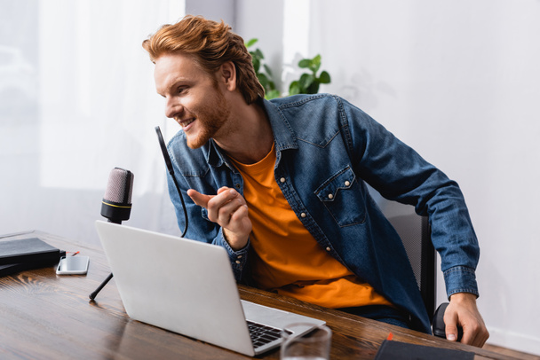 redhead broadcaster pointing with finger while speaking in microphone near laptop - Photo, Image