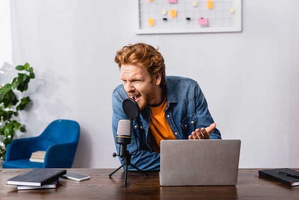 angry redhead announcer gesturing while screaming in microphone near laptop - Photo, Image